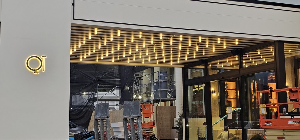 Site progress photograph of illuminated pendant lights under the QT porte cochère, with scaffolding and access equipment visible at the entrance.